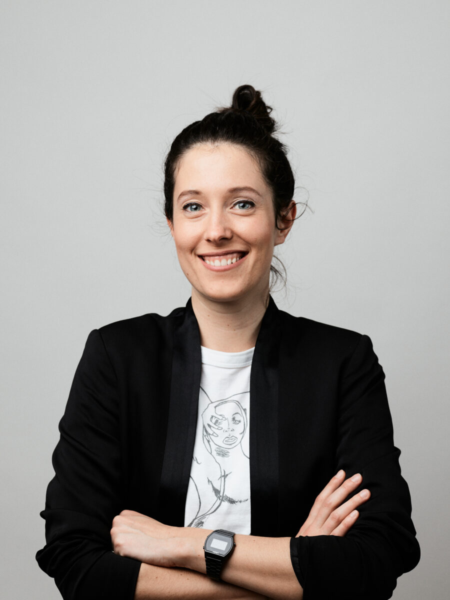 Portrait of young lady – black hair, blue eyes, wearing a white shirt and a black jacket –  in front of grey wall