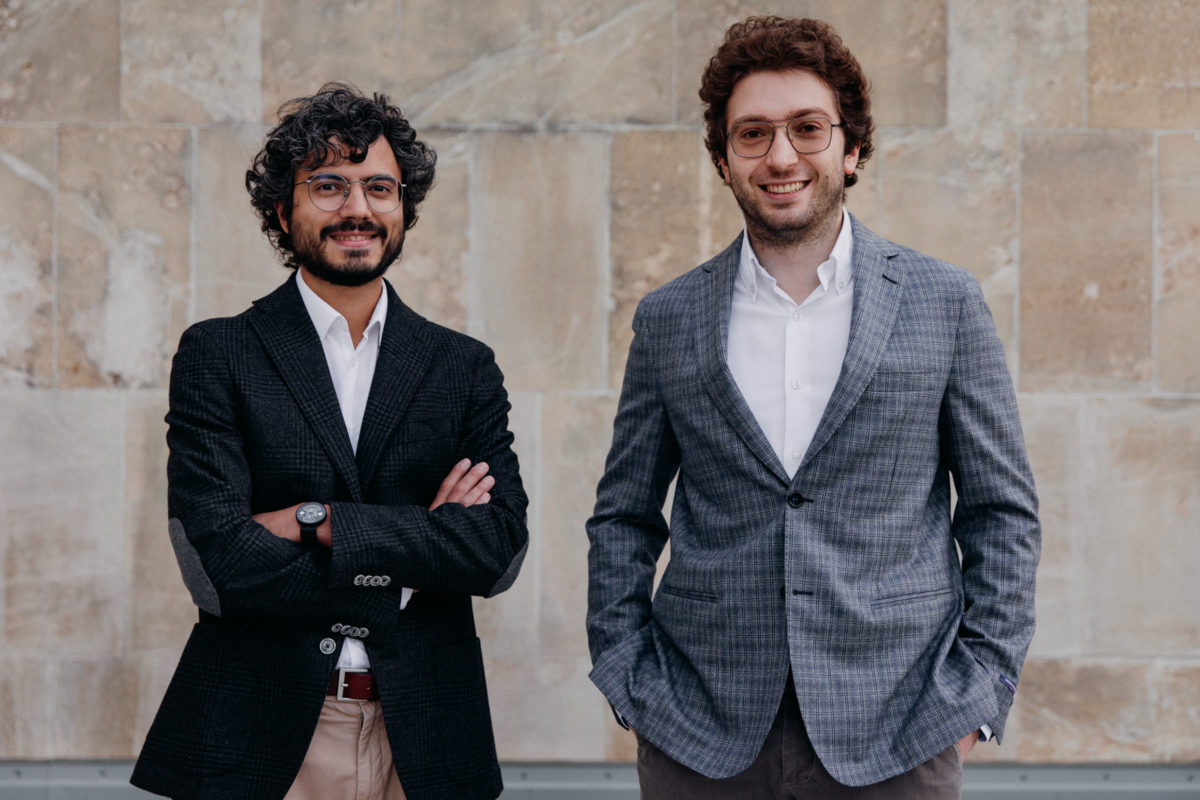 two young men, the founders, with dark hair and jackets standing in front of a wall, smiling