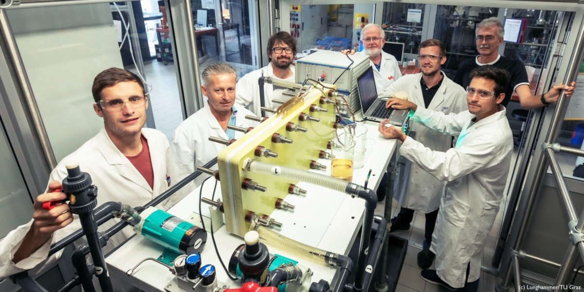 Team of people stranding in a laboratory in front of a battery wearing white coats 