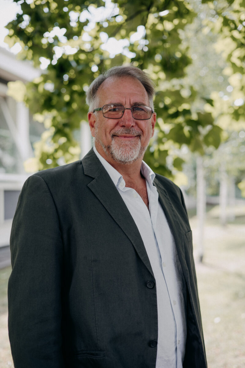 Man with beard and glasses wearing a jacket standing in front of a tree