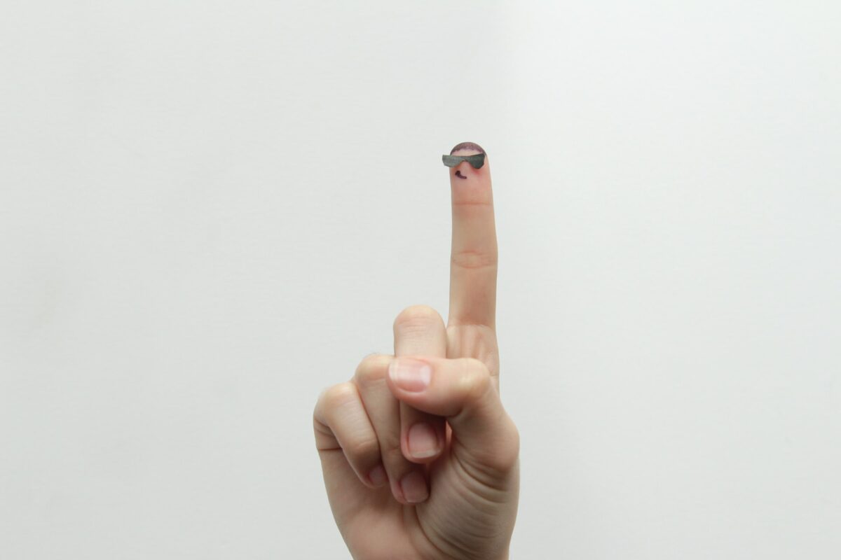 hand with raised finger on white background. Fingertip is wearing glasses and smiling. 