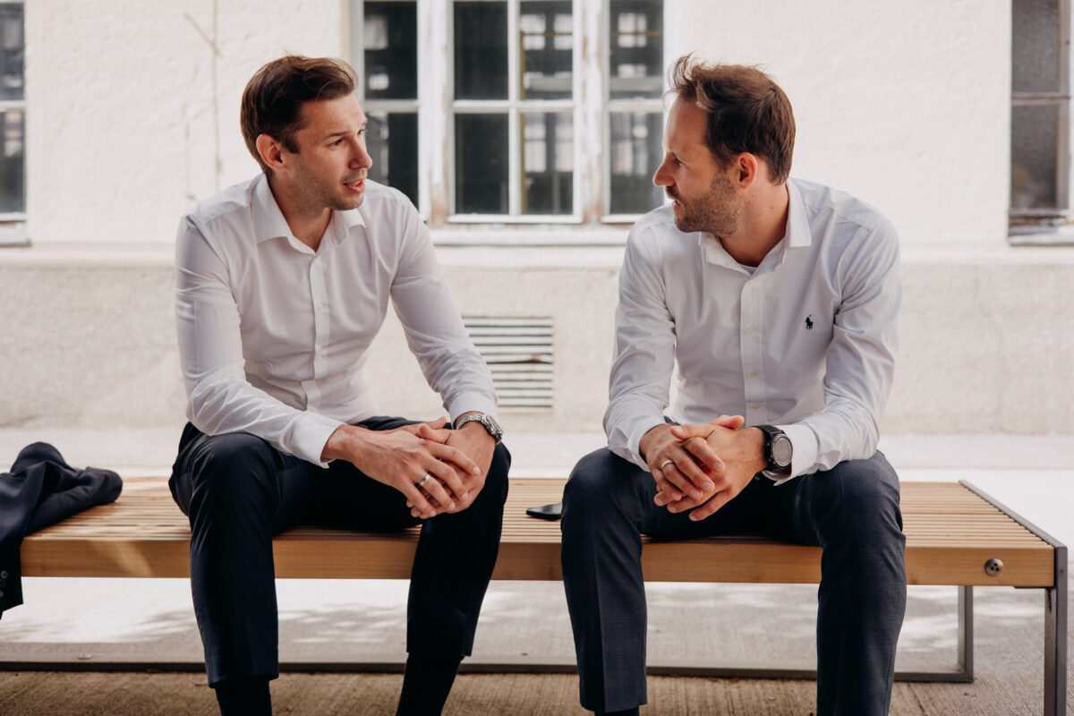 Two men in white shirts sitting on a bench in front of a building