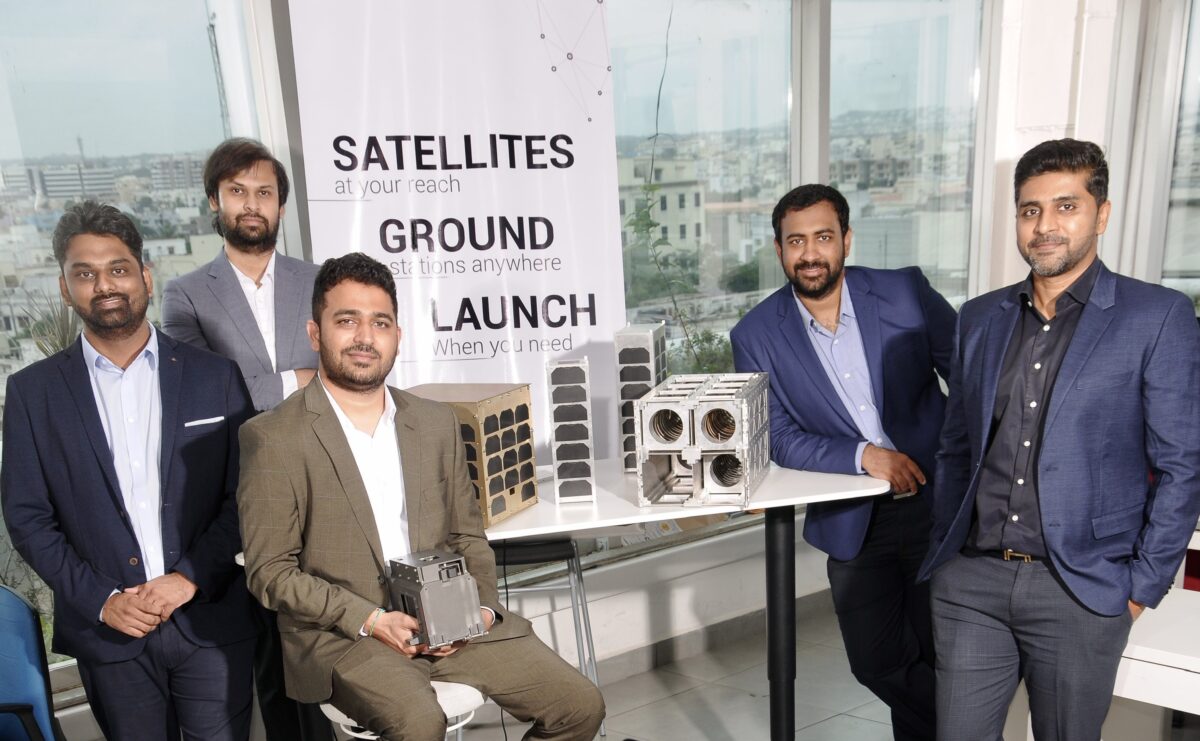 5 men in a room sitting and standing at a table in front of a roll up holding samples 